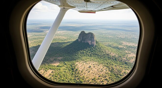 Looking out the window of a light aircraft flying over a remote wilderness, with an aerial view of a large inselberg mountain surrounded by an endless forest on a sunny day