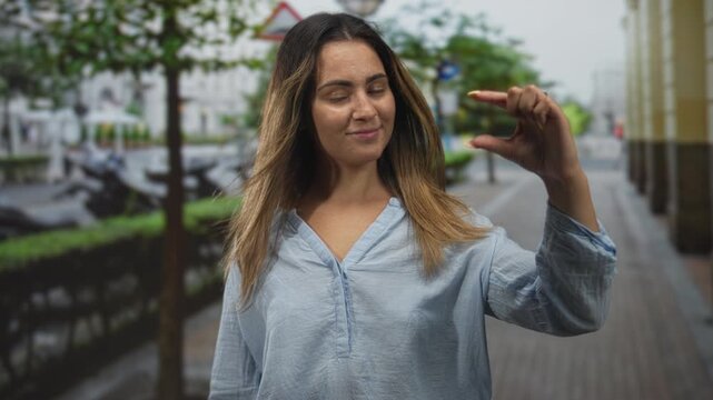 Woman in light blue blouse pinching a tiny gap between fingers on a wet city street sidewalk with scooters and trees visible; confidence measurement.