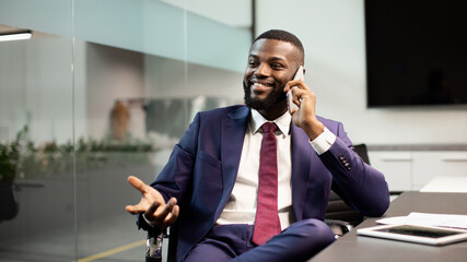 Attractive millennial african american man boss sitting at workdesk at office, having phone conversation with personal assistant or business partner, looking at copy space, gesturing and smiling
