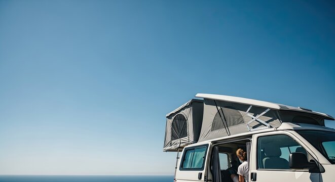 A woman relaxes inside her camper van with a pop-up roof tent, enjoying a scenic ocean view on a sunny day during a coastal road trip adventure