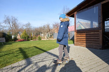 Teen skateboarding in sunny garden wearing winter coat and beanie