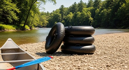 A stack of black inflatable tubes and a canoe with a paddle rest on a rocky riverbank, ready for a fun summer day of floating and adventure in the peaceful wilderness