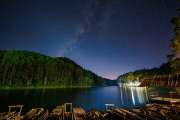The Milky Way and a starry night sky over a calm lake with pine forest and wooden rafts in the...