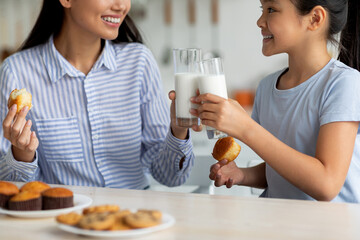 In a bright kitchen, a young girl and her mother smile as they enjoy milk together. The girl holds a glass of milk while the mother eats a treat, creating a joyful moment of connection.