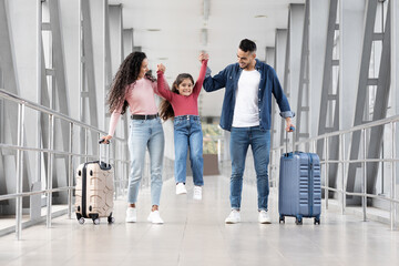 A family is walking through the airport. A mother and father lift their daughter happily while pulling their suitcases. The setting is bright and modern, full of excitement for travel.