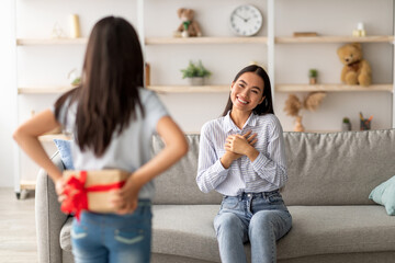 A young girl hides a wrapped gift box behind her back, smiling as she wishes her surprised mother a happy Mother's Day. They are enjoying a warm family celebration in their cozy living room.