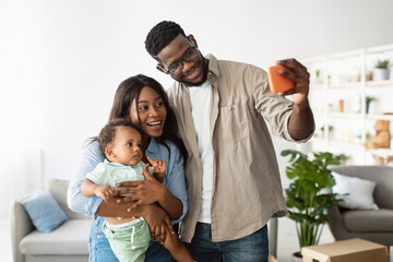 This joyful family captures a memorable moment in their new home. A man holds a smartphone while standing with his wife and son, celebrating their recent move in a bright living space.