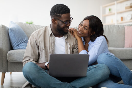Cheerful black couple sits together on the floor near their couch, using a laptop to browse for new furniture while sharing joyful moments at home.
