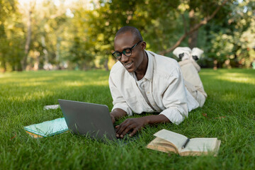 A person is lying on the grass in a park, focused on a laptop. Nearby are books and notebooks, while sunlight filters through the trees, creating a serene atmosphere.