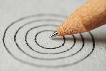 Close-up of a pencil drawing a target on a piece of paper. A wooden pencil is shown drawing a series of concentric circles on a textured paper surface.