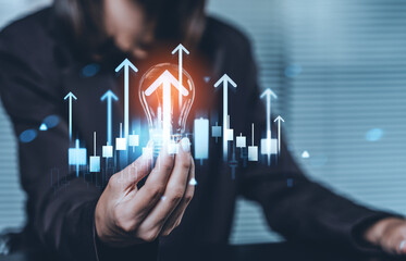 Businessperson holding glowing lightbulb with upward arrows and candlestick charts on a futuristic background symbolizing innovation and financial growth concept.