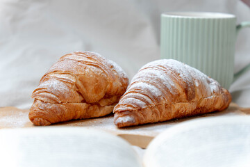 Freshly baked croissants with powdered sugar and a green coffee mug, set against a cozy bed background.