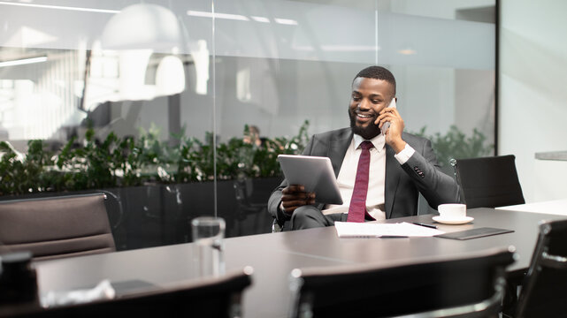 Happy young black guy in suit entrepreneur sittig at desk at office, using modern digital tablet and talking on phone, panorama with copy space. Modern technologies and business concept