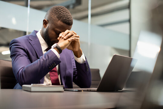 Stressed african american young businessman in suit having burnout, sitting in his office in front of laptop, leaning on his hands, suffering from financial crisis during COVID-19 pandemic, copy space
