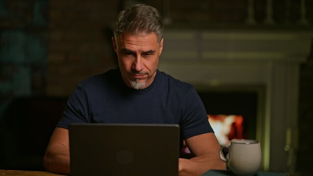 Mid adult man working with laptop computer in living room with fireplace in winter. Businessman managing business in home office, banking online, remote working, browsing internet. - Powered by Adobe