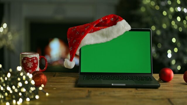 Laptop computer in Santa hat at Christmas. Decorated room with Christmas tree and lights. Cosy winter home with fireplace. Blank green screen for web shop offer and holiday advertisement. - Powered by Adobe