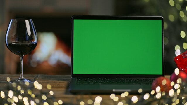 Glass of red wine with laptop computer on table in Christmas decorated room. Christmas tree and lights in winter home with fireplace. Green screen for web shop ads and vinery holiday offer.