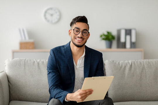 A cheerful arab male psychologist sits on a sofa in a clinic office, smiling at the camera while taking notes. The setting is calm and professional, ideal for therapy sessions with clients.