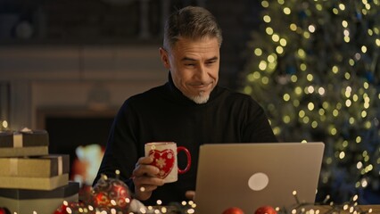 Happy middle aged man with laptop computer at home, sitting at desk in Christmas decorated cosy room. Fireplace, Christmas tree in background. Buying gifts in web shop, searching presents online.