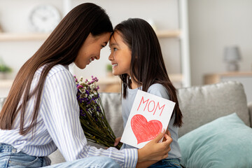 A young girl joyfully greets her mother with a bouquet of flowers and a handmade card. © Prostock-studio