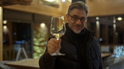 Expert drinking, tasting, toasting glass of red wine. Middle aged man at home wearing turtleneck pullover and warm vest. Eyeglasses and gray hair. Satisfied and smiling. Autumn or winter clothing.