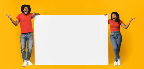 A cheerful couple poses with a large blank sign. They are in casual clothing, standing in front of a vibrant yellow backdrop. The scene captures their fun and lively spirit.