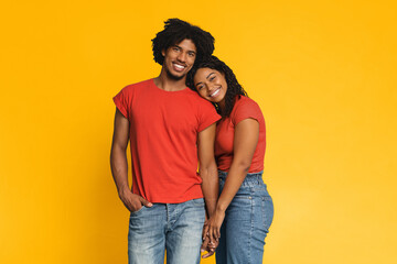 A couple poses together, smiling and holding hands. They wear matching red shirts and comfortable jeans. The background is a vibrant yellow, adding to the cheerful atmosphere of the scene.