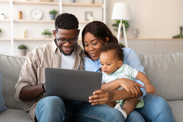 Happy parents share a funny educational video on a laptop with their young son while relaxing on the sofa at home. The family is clearly excited and engaged in this joyful moment.