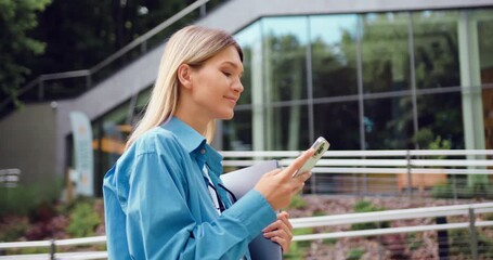 Side view of young Caucasian businesswoman in blue shirt using smartphone and holding laptop while walking in front of modern glass building at city park. Business and people concept. - Powered by Adobe