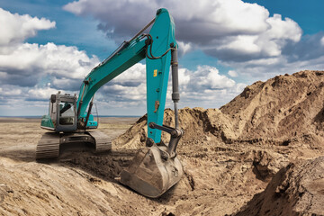 A large excavator works on a construction site, digging through sandy soil with a backdrop of clouds during the day. Dust rises as the machine operates