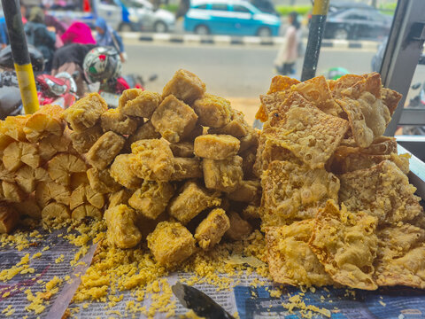 Pile of Indonesian street food Gorengan. Deep fried tofu and tempeh fritters displayed on a vendor cart with a blurry street background.