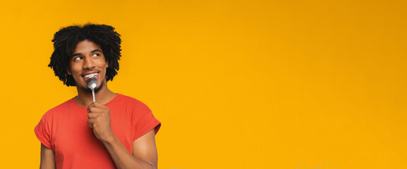 A young man with curly hair wears a red shirt and grins while holding a spoon near his mouth. The bright yellow background adds a cheerful atmosphere to the scene.