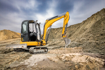 A yellow excavator operates at a construction site, digging into the earth and moving dirt under a cloudy sky in the background