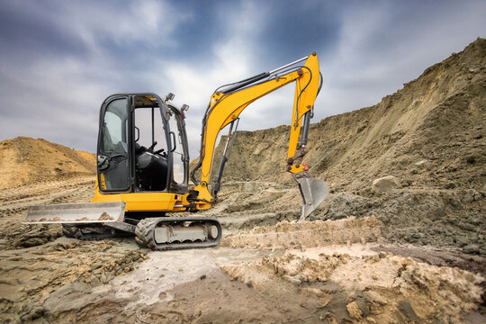 A yellow excavator operates at a construction site, digging into the earth and moving dirt under a cloudy sky in the background