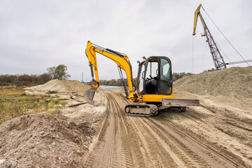 Obraz premium Construction equipment digs and moves earth along a riverbank, surrounded by a cloudy sky and rugged terrain on a working site