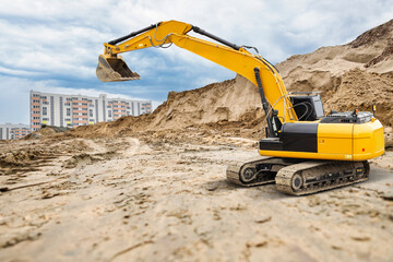 A powerful crawler excavator is seen digging on a sandy construction site, surrounded by tall residential buildings under a cloudy sky