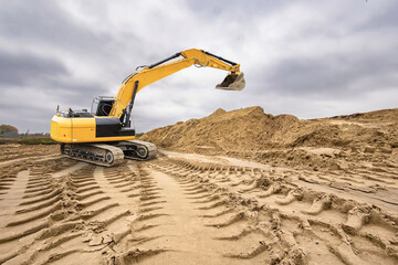 A yellow excavator digs into the sandy ground at a construction site with piles of earth under a...