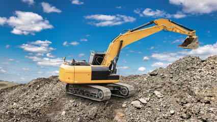 An excavator is moving dirt at a construction site with a clear blue sky and scattered clouds overhead. Piles of gravel and rocks are visible around the machine