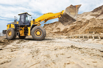 Obraz premium A large yellow A wheeled bulldozer or loader is moving sand at a construction site, working efficiently under a partly cloudy sky with a city backdrop