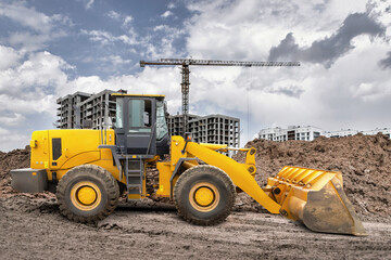 Obraz premium A yellow loader is working at a construction site, moving dirt while cranes and new buildings rise in the background on a cloudy day