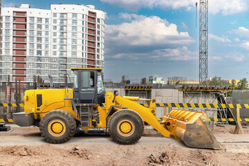 A yellow loader operates on a construction site, surrounded by new buildings and a bright blue sky, showing activity in urban development