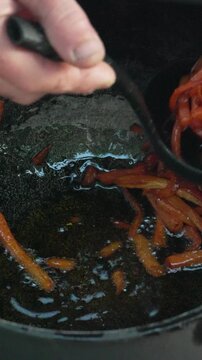 Carrots, onions, frying. Hand scooping caramelized vegetables from hot oil in a pan, culinary preparation. Vertical video.