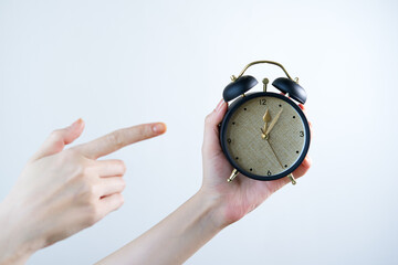 Female hand holding an alarm clock and pointing her finger at it against gray background.