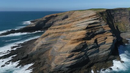 Coastal headland rises sharply above surf, exposed rock layers forming horizontal bands across the cliff face. - Powered by Adobe