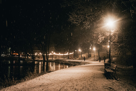 Snow-covered pathway illuminated by street lamps at night, with gentle snowfall creating a serene atmosphere, inviting peaceful winter strolls along the riverbank