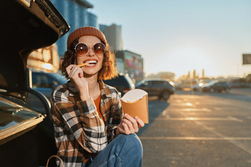 woman eats fries by car trunk in parking lot, smile and sunglasses catching sunlight as she sits in...
