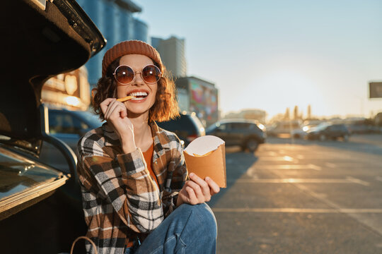 woman eats fries by car trunk in parking lot, smile and sunglasses catching sunlight as she sits in beanie and plaid jacket at golden hour authenticity, candid lifestyle, golden hour glow, mindful