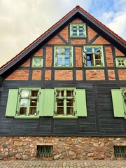 Historic half-timber house with green wooden windows and shutters, black beams, brick walls, and a stone foundation. Traditional European architecture.