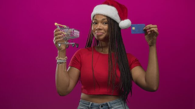 Woman wearing santa hat holding a mini shopping cart and credit card in studio with magenta backdrop; holiday shopping joy.