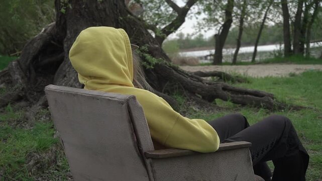 girl yellow hoodie sits motionless old vintage chair, hiding hands pockets admiring beauty local nature outside city, against backdrop large thick old willow tree beach river